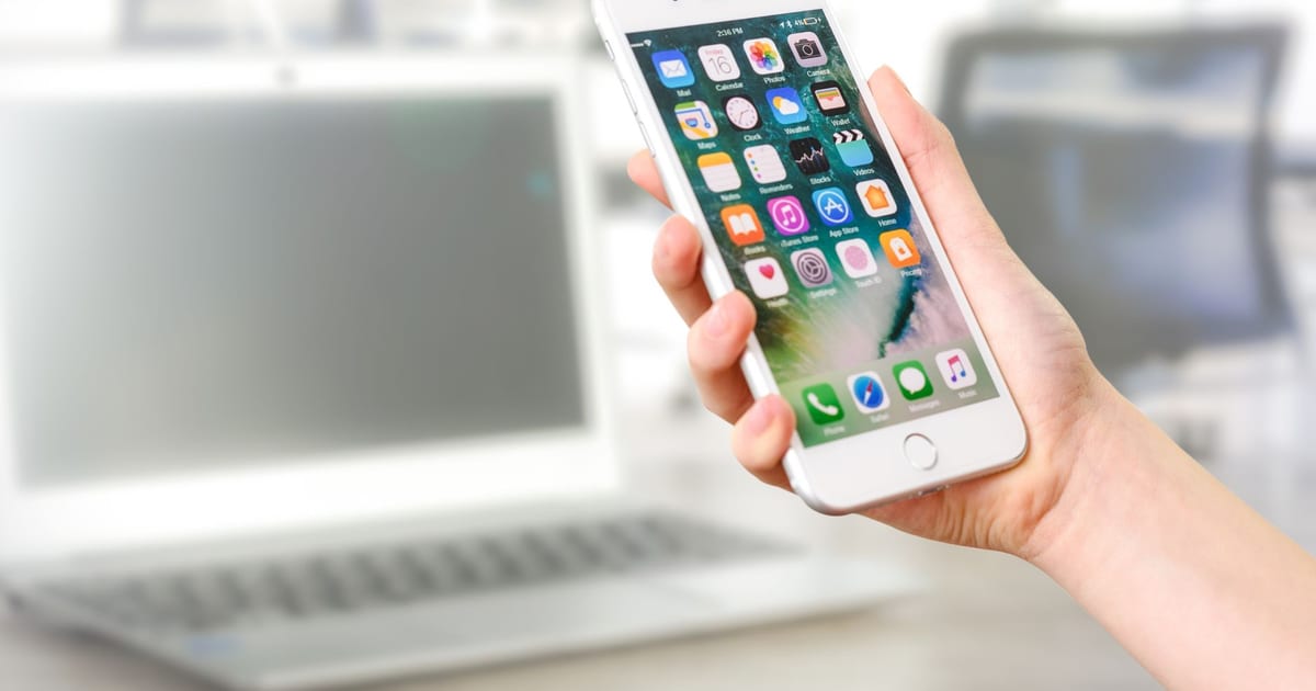 Hand holding a smartphone displaying apps, with a laptop blurred behind